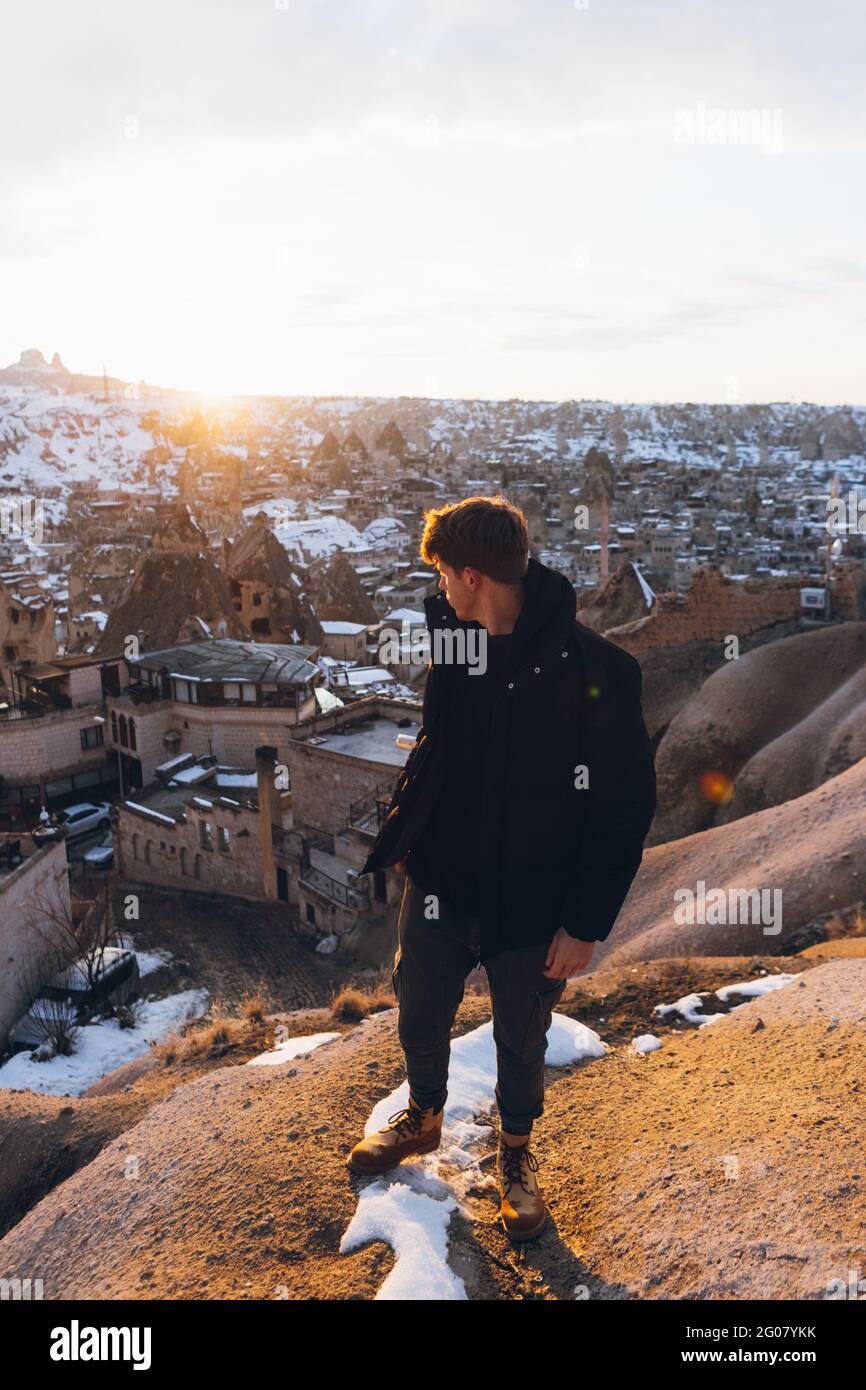 From above man in warm clothes looking over shoulder while standing on snowy hill against famous small town with ancient cave houses in valley during Stock Photo
