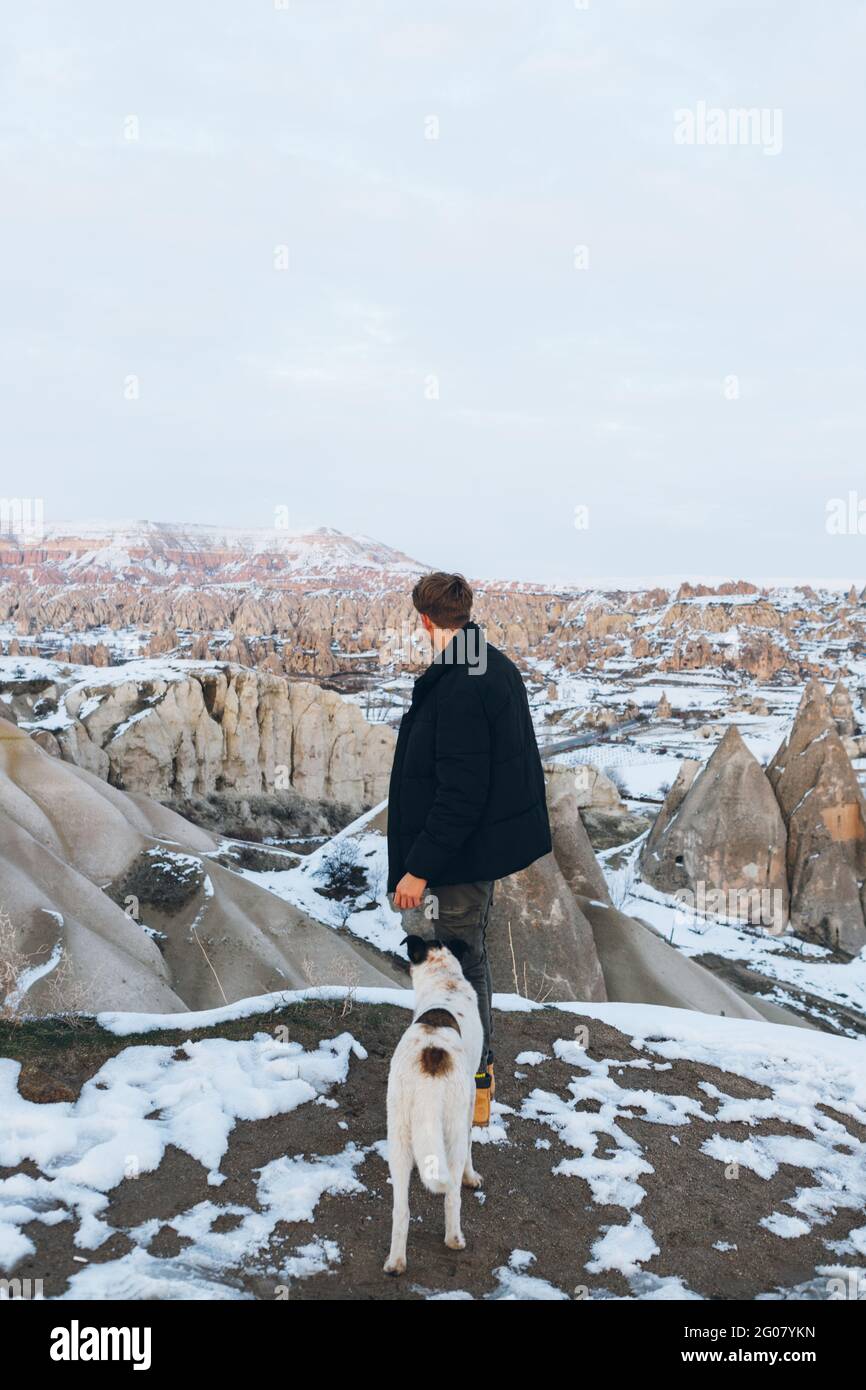 Back view of unrecognizable man in warm clothes with loyal dog in snowy hill against misty mountains in overcast weather in Turkey Stock Photo