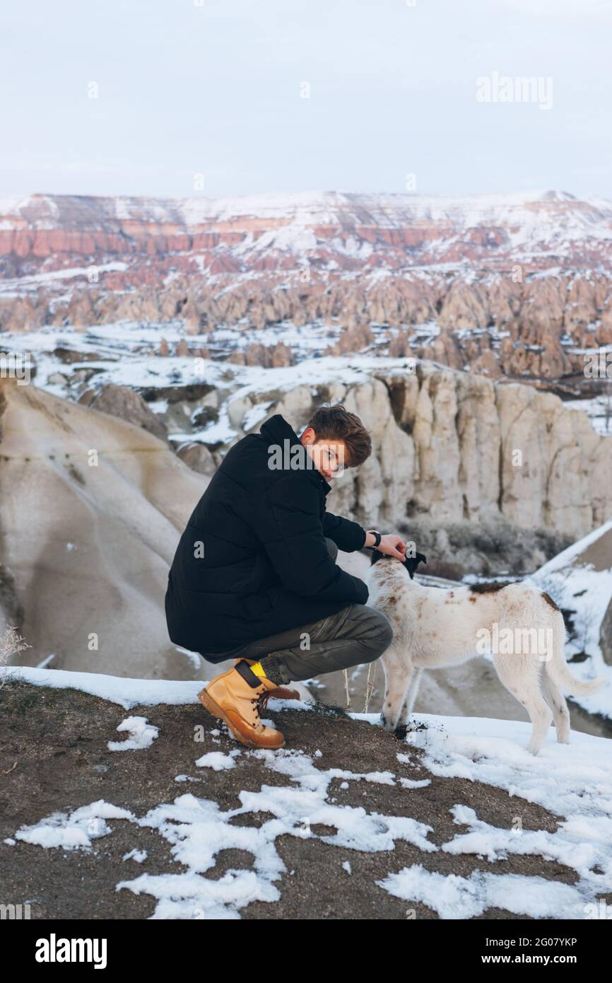 Young man squatted down in warm clothes caressing dog in snowy hill against misty mountains in overcast weather in Turkey Stock Photo