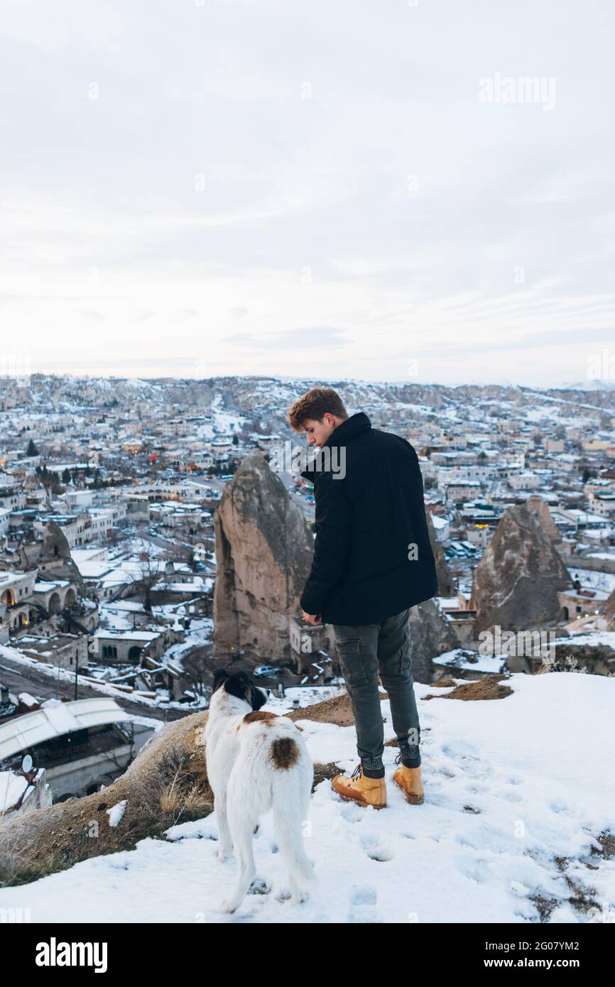 Back view of a young man in warm clothes standing with loyal dog on hill against small ancient cave houses in snowy valley at dusk in Turkey Stock Photo