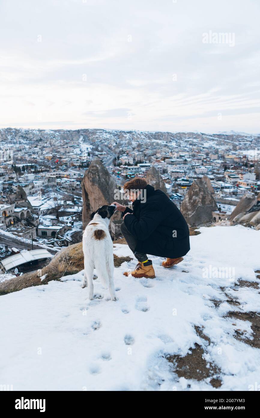 Back view of a young man squatted down in warm clothes with loyal dog on hill against small ancient cave houses in snowy valley at dusk in Turkey Stock Photo