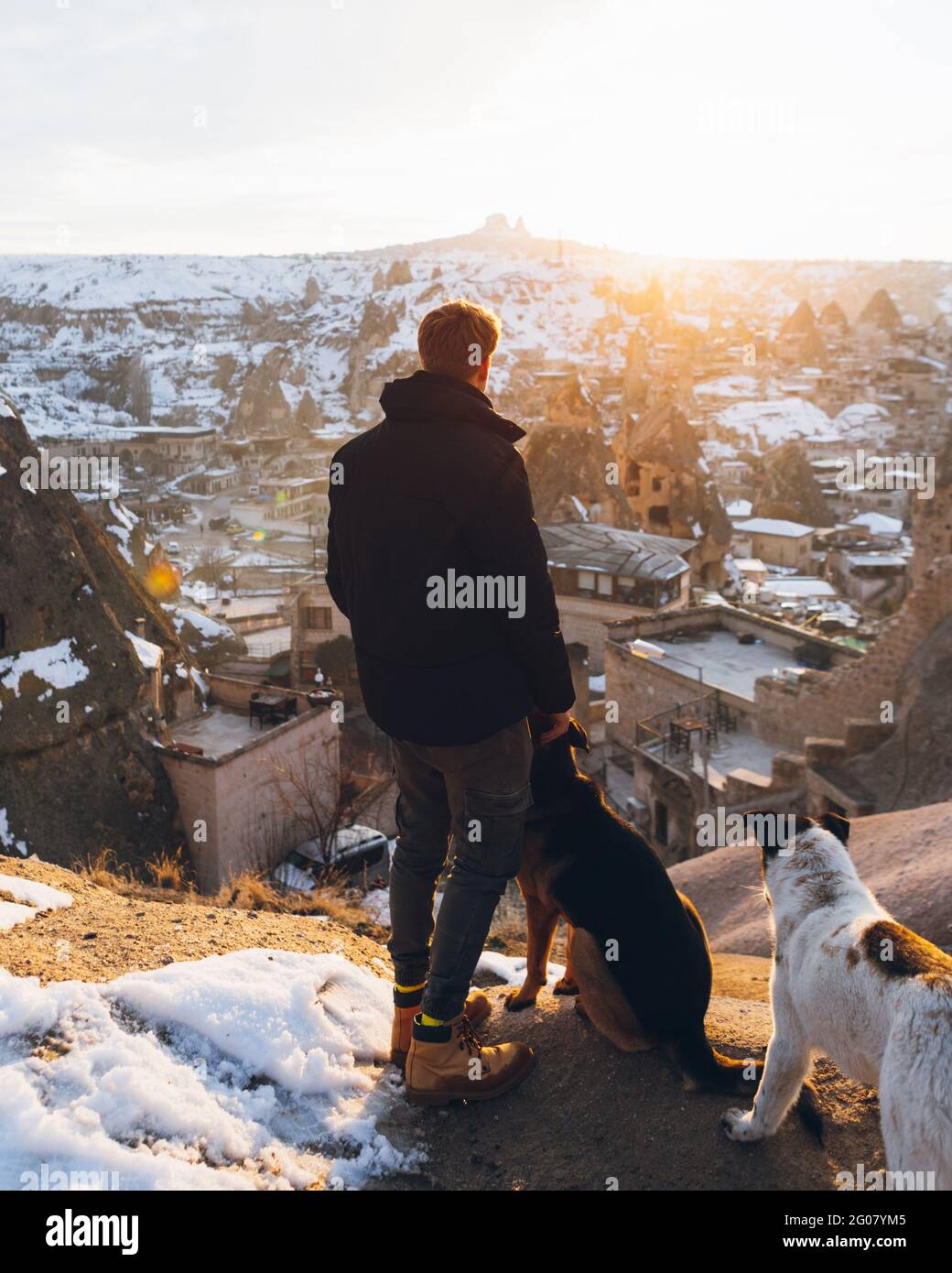 Back view of a young unrecognizable man in warm clothes standing caressing dogs on hill against small ancient cave houses in snowy valley at dusk in T Stock Photo