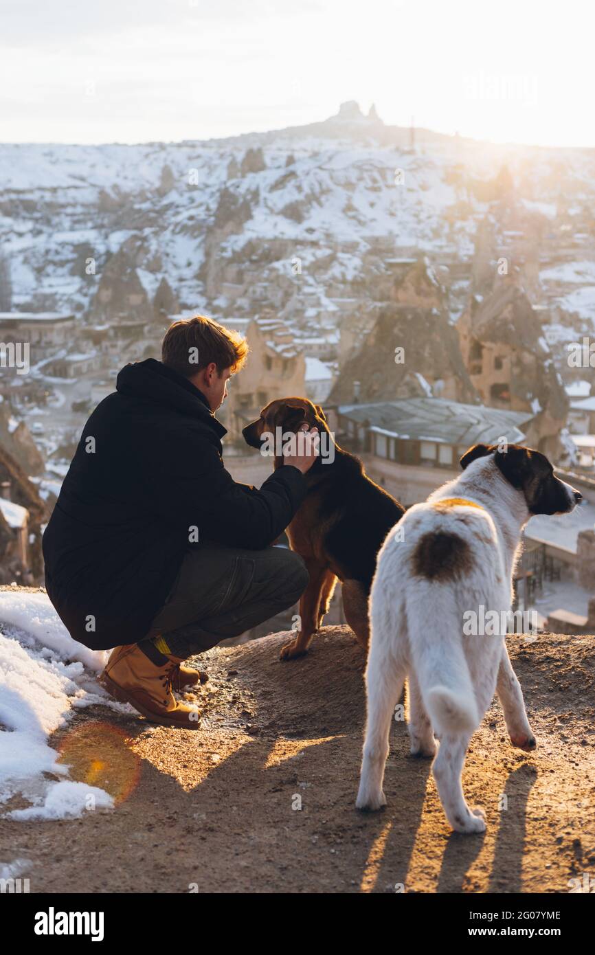 Young unrecognizable squatted down man in warm clothes caressing dogs on hill against small ancient cave houses in snowy valley at dusk in Turkey Stock Photo