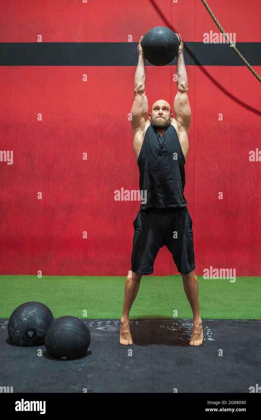 Full length strong male athlete lifting heavy ball in raised arms while