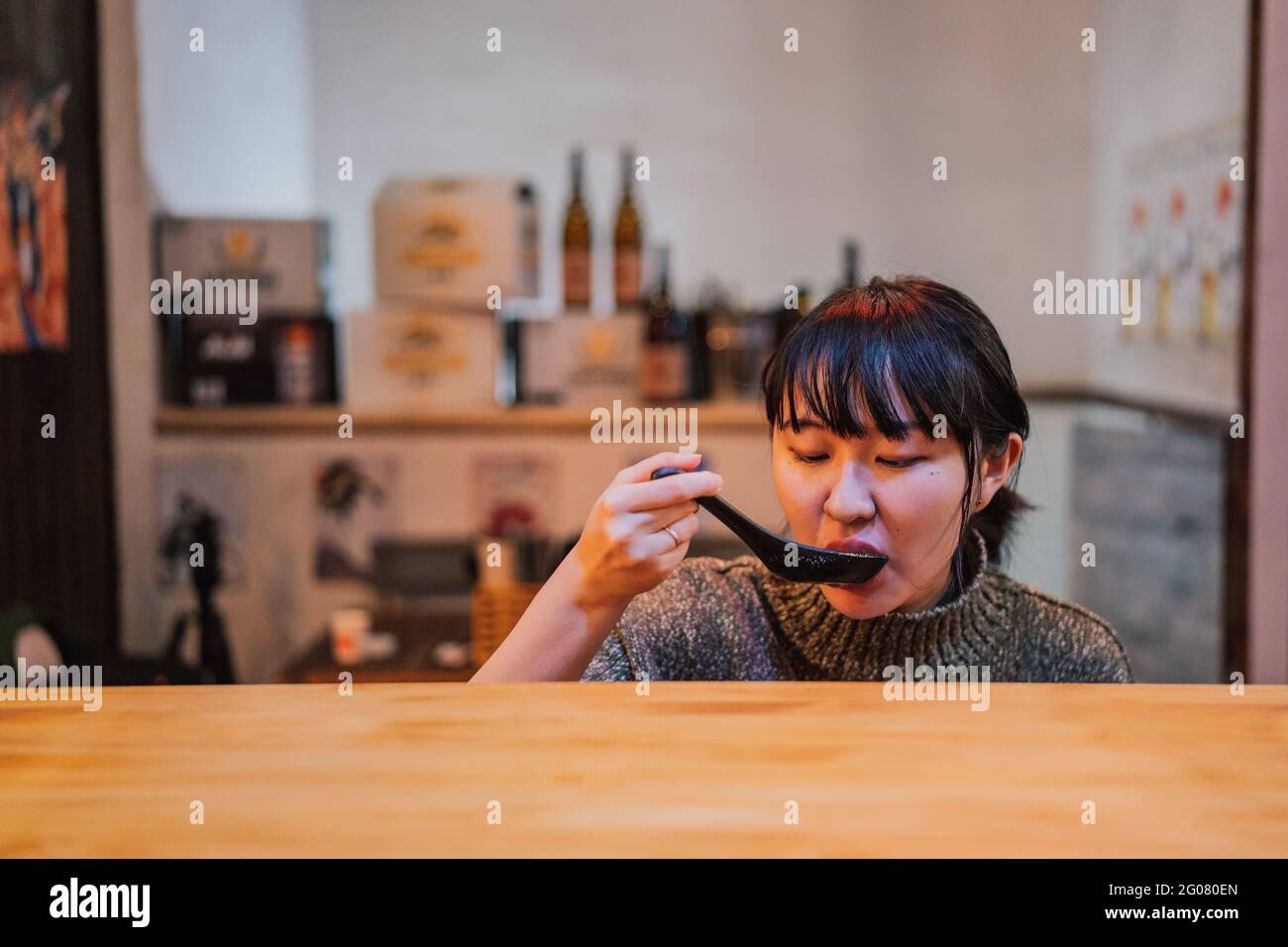 Young Asian Woman in sweater eating ramen with spoon at wooden counter in cafe Stock Photo