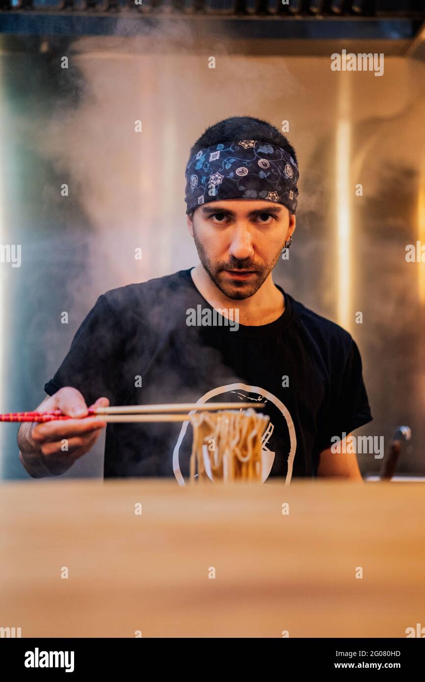 Young bearded male chef in bandana looking at camera while holding noodle with chopsticks in ramen bar Stock Photo