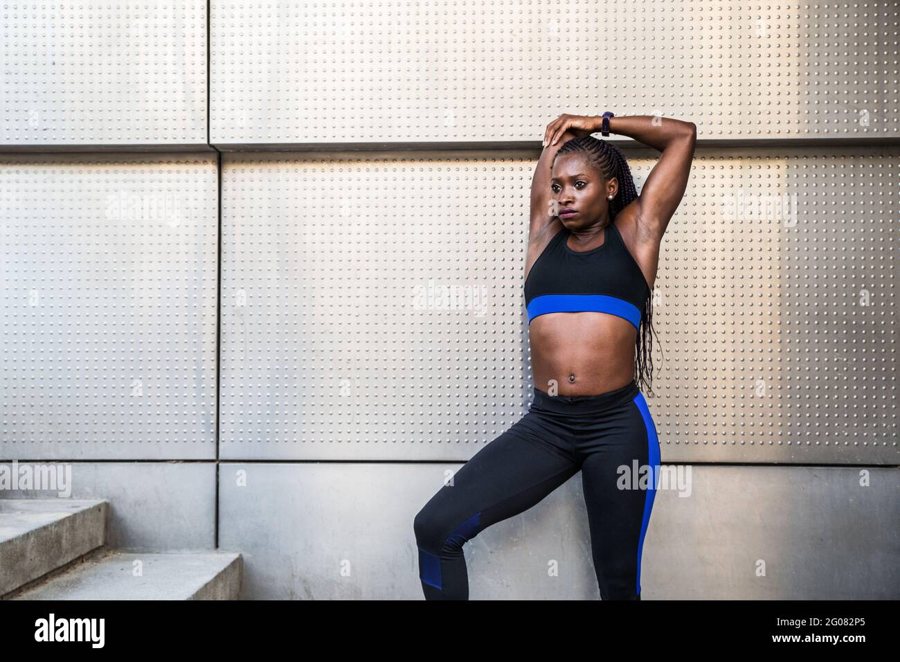 Pretty black female in sportswear keeping eyes closed and bending aside while doing warming up exercises near wall of modern building on city street Stock Photo