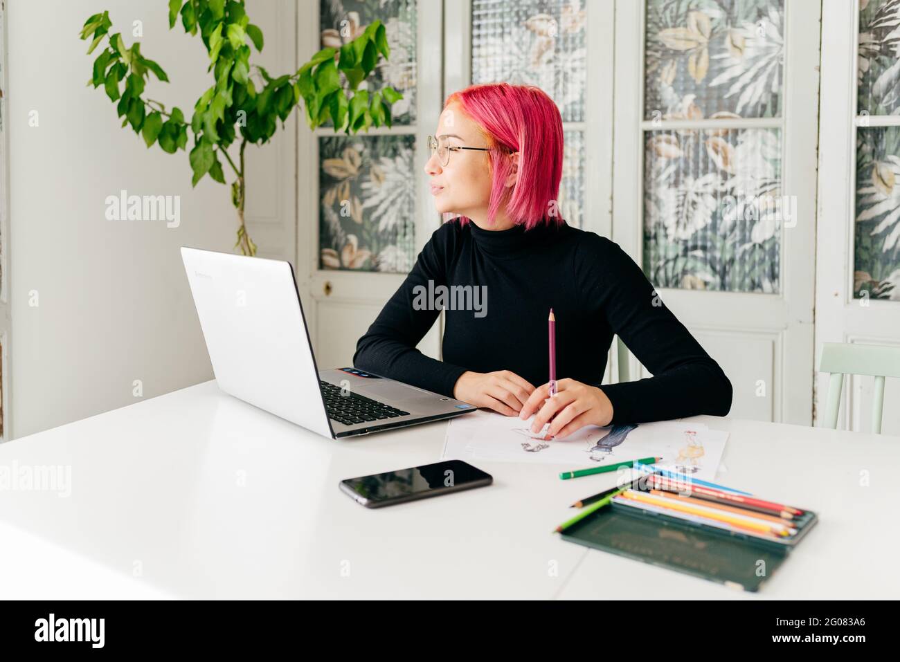 Young creative female fashion designer in casual outfit and glasses looking away pensively while sitting at table with gadgets and drawing sketch with Stock Photo