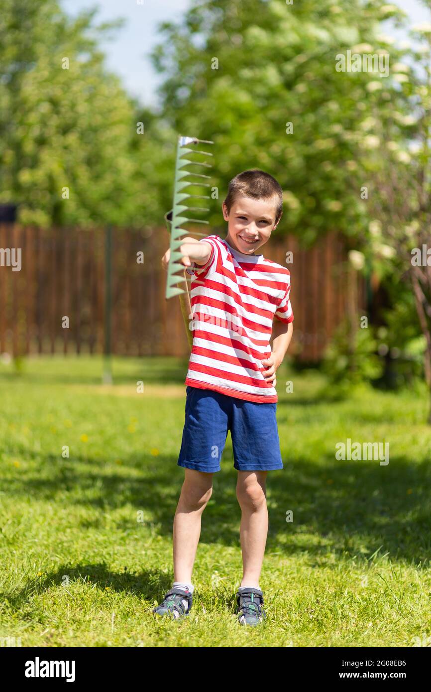 A little cheerful boy stands and holds a rake in his hand, ready to ...