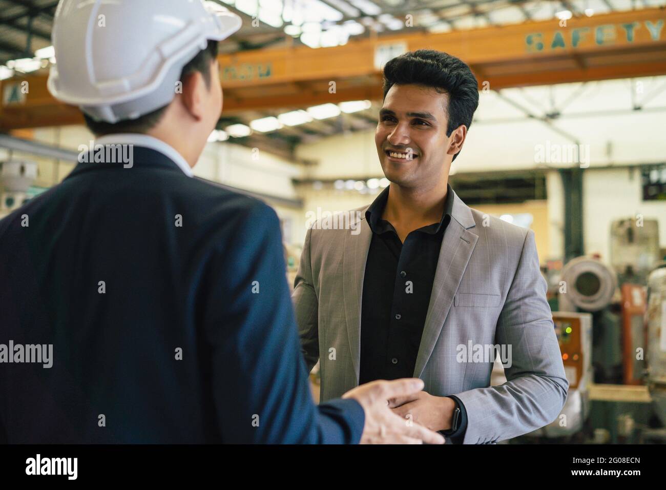 30s young Asian businessman in formal suit and hard hat showing foreign business partner in factory background. Business partnership and team work success concept Stock Photo