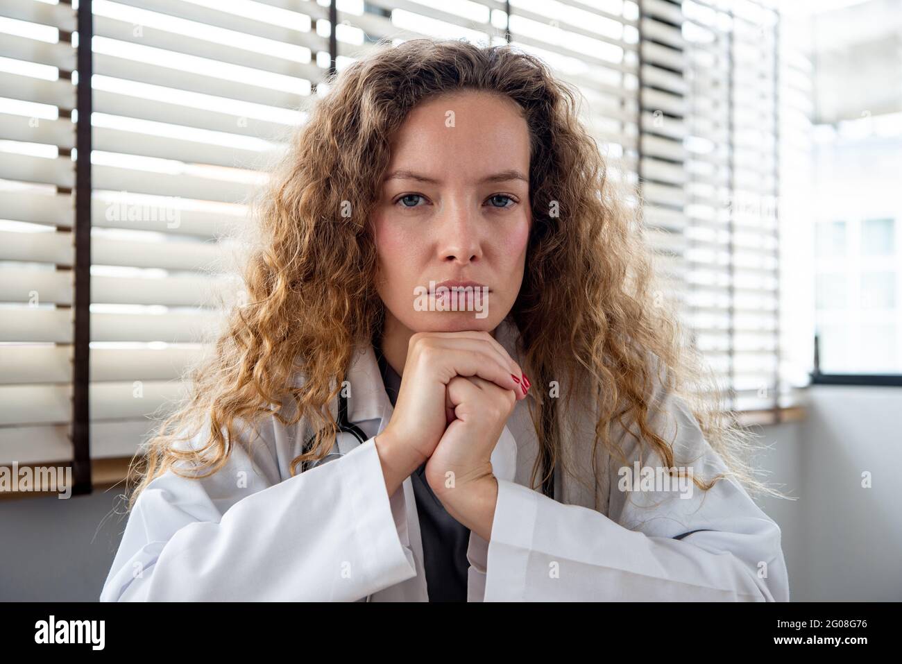Serious female doctor carefully listening and keeping hands under chin while making medical conference video call from hospital, telehealth concept Stock Photo
