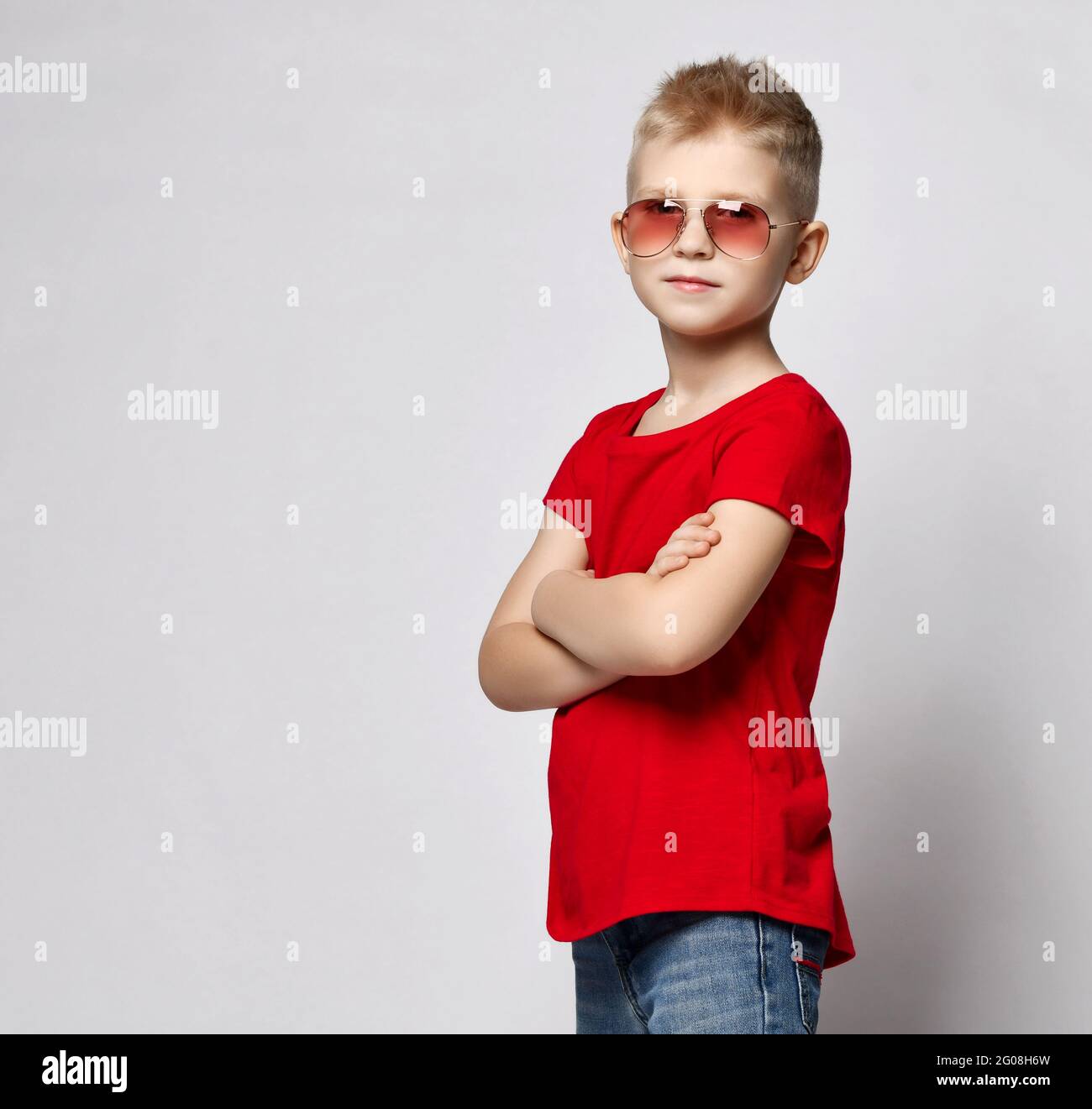 Stylish kid boy child in red t-shirt, jeans and sunglasses standing with hands crossed looking at camera Stock Photo