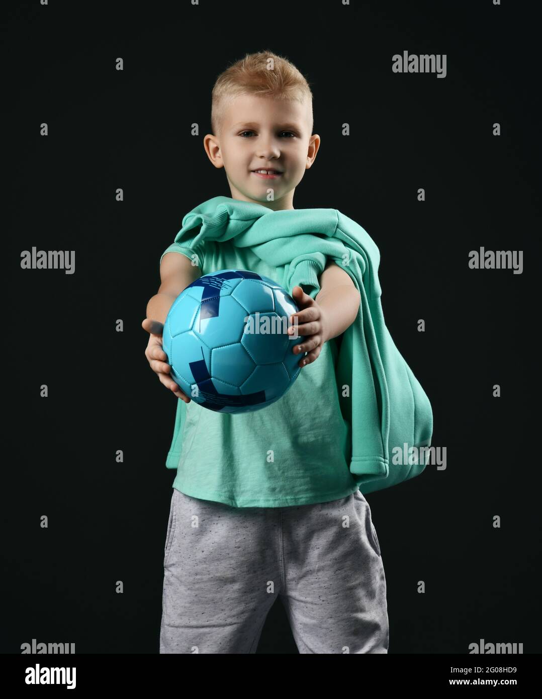 Smiling blond boy child in sports green t-shirt and trousers standing holding soccer ball in hands Stock Photo