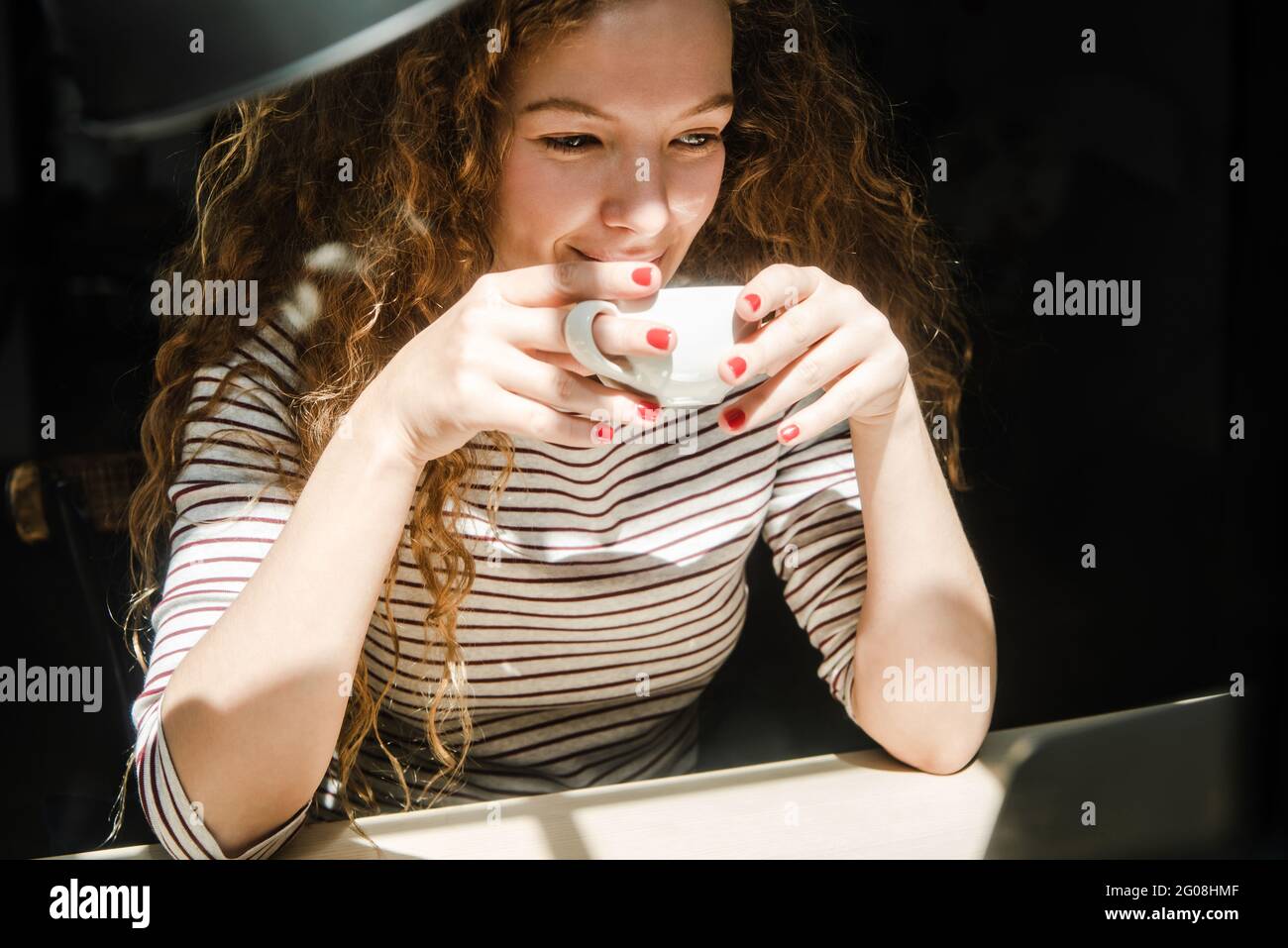 Happy young Caucasian woman drinking coffee while watching video on laptop computer at home Stock Photo
