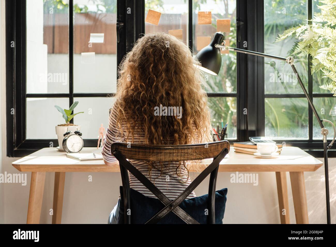 Back view of Caucasian woman sitting and working near the window at home in the morning Stock Photo