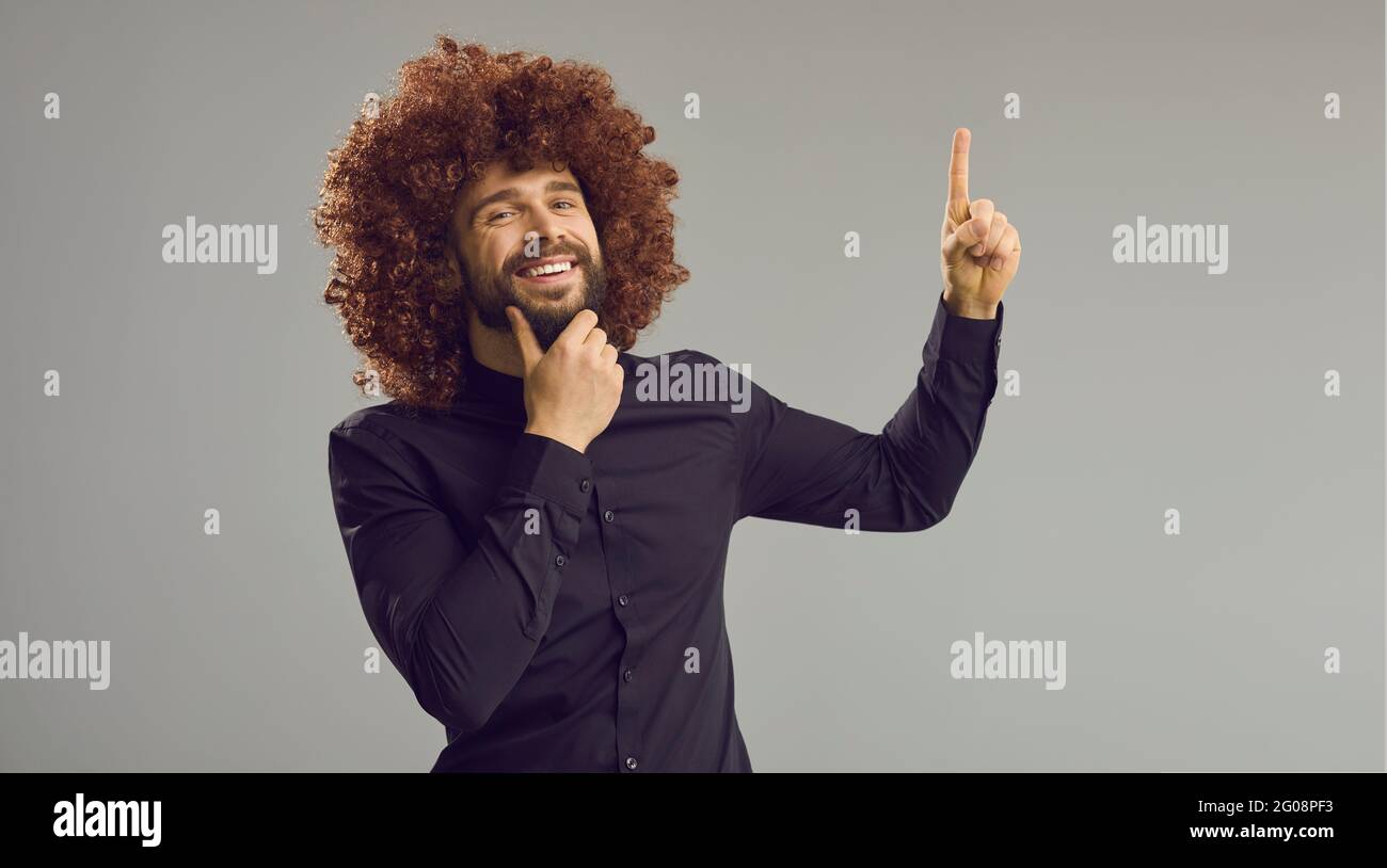 Funny millennial man in a funny curly wig standing on a gray background thinks and has an idea. Stock Photo
