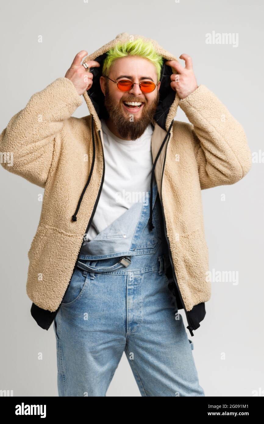 Happy hipster model with beard and bright hair, looking at camera and laughing loud out, putting on hood, expressing positive emotions. Indoor studio Stock Photo