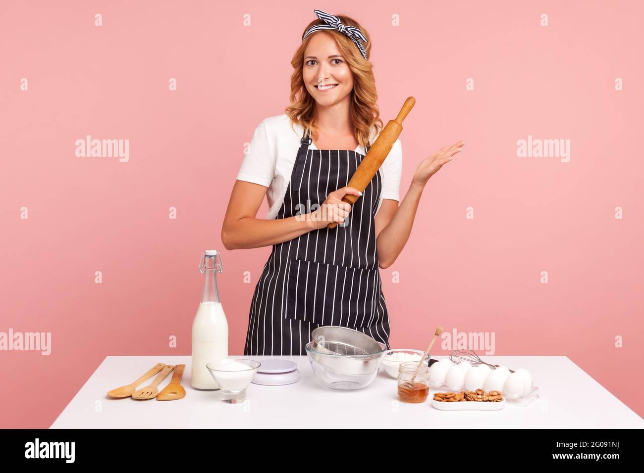 Happy attractive young adult woman housewife baker wear apron holding rolling pin in hands, baking pastry, cooking cake biscuit doing bakery. Indoor s Stock Photo