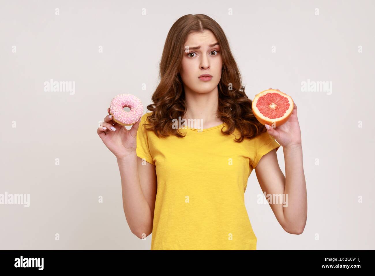 Sad teenager girl holding healthy fruits and high-calorie dessert, confused wavy-haired female in T-shirt choosing between grapefruit and donut. Indoo Stock Photo