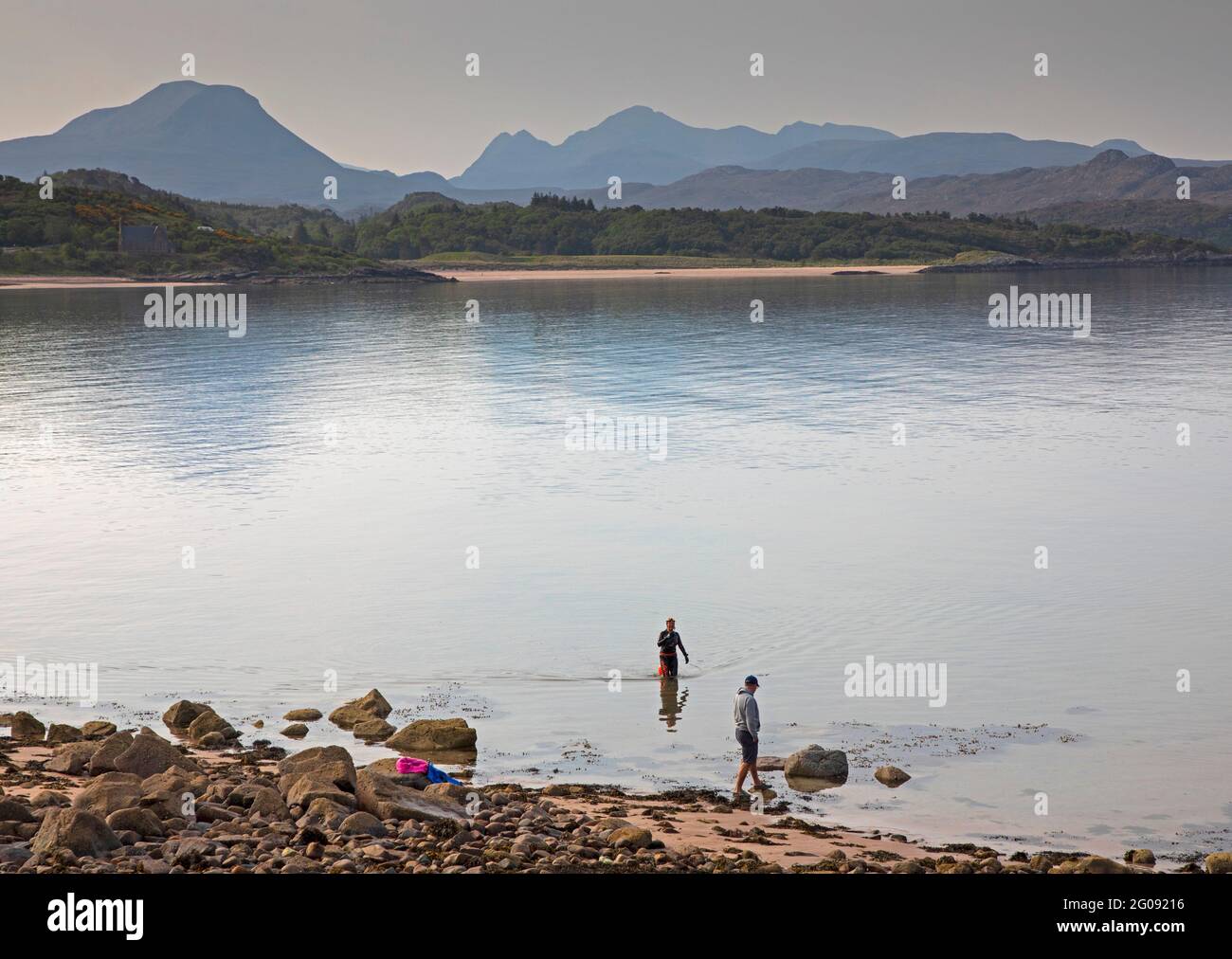 Strath By, Loch Gairloch, Wester Ross, Scotland, UK weather, 2nd June 2021. Hazy sunshine in morning, temprature  of 12 degrees centigrade with 21 degrees expected later, currently almost midge free.. Pictured: Female Cold water swimmer in wet suit leaving the loch walking towards her partner after a cool dip, with the Torridon mountains in the background. Credit: Arch White/Alamy Live News. Stock Photo