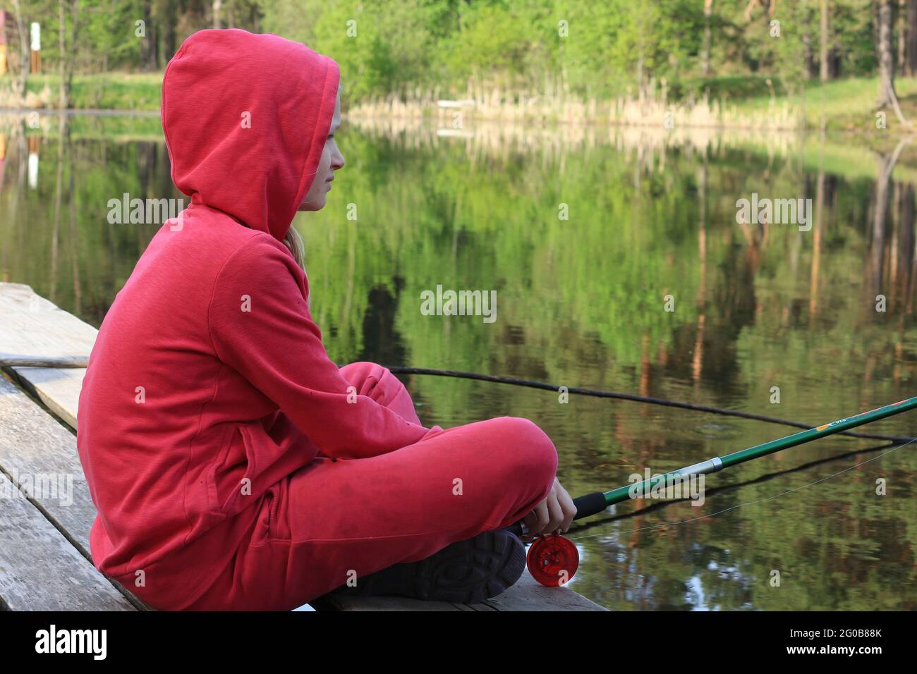 A girl in a red tracksuit is fishing in a forest lake sitting on a wooden