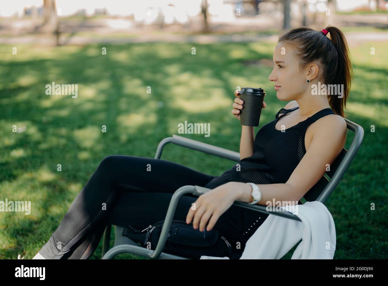 Thoughtful sporty young woman with pony tail has coffee break after training poses in comfortable chair with hot drink in park against green grass Stock Photo