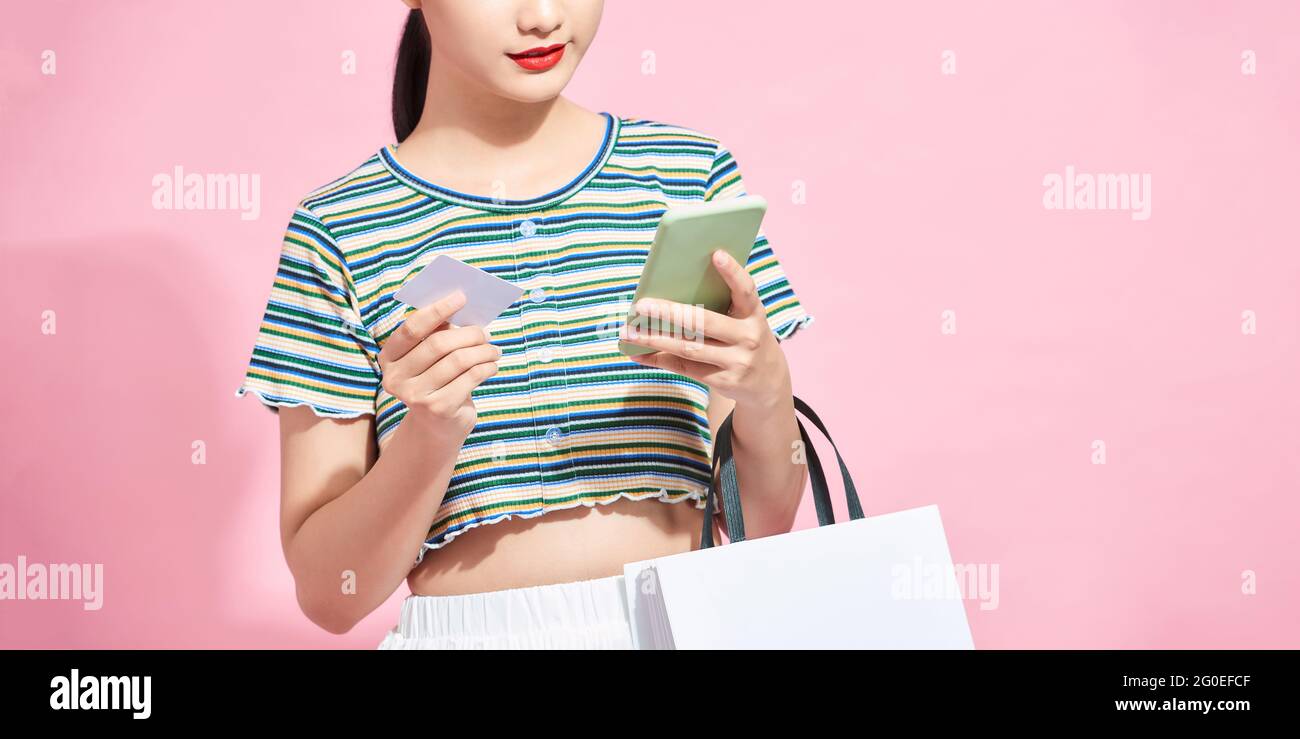 young smiling beautiful Asian woman making online payment through mobile phone via credit card while carrying white shopping bags in isolated pink bac Stock Photo