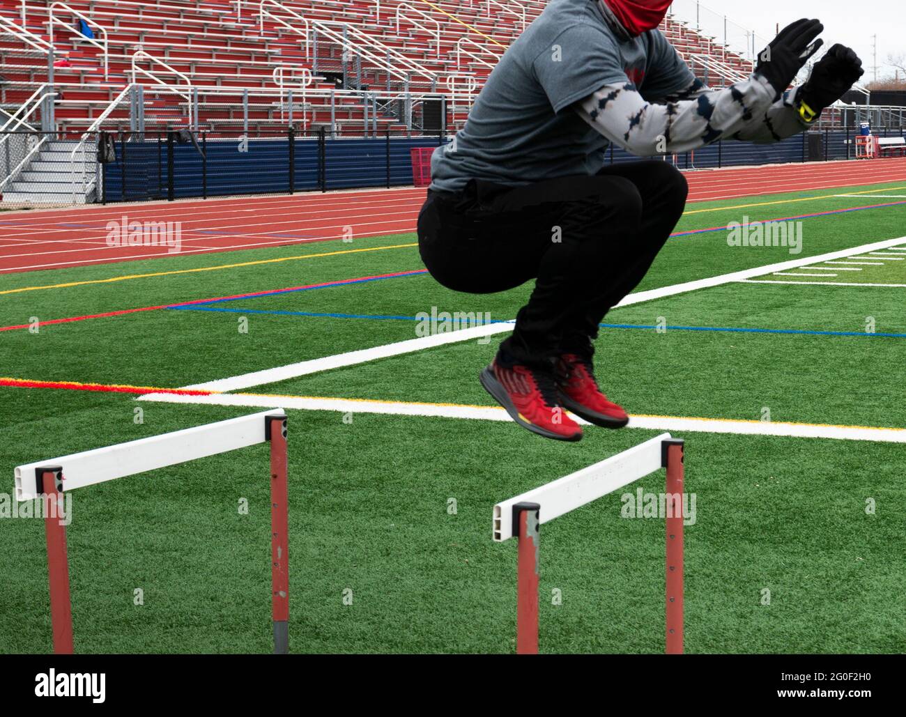 A male high school track runner is jumping over hurdles on a green turf field during track and field practice in the winter. Stock Photo