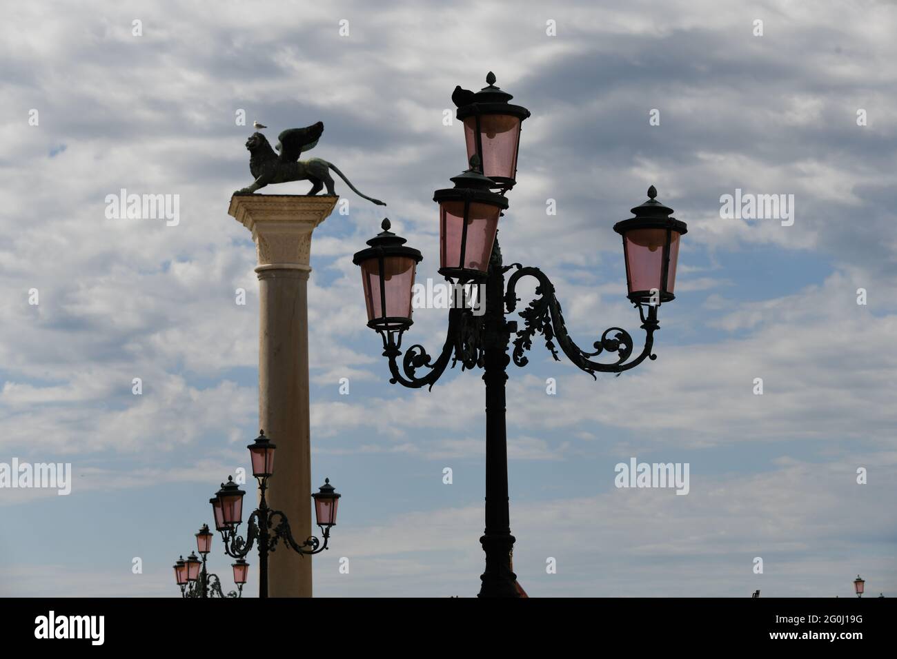 Vierarmige Strassenlaterne am Eingang zum Markusplatz in Venedig gegen den bewölkten Himmel, im Hintergrund das Wahrzeichen der Stadt, der Löwe Stock Photo