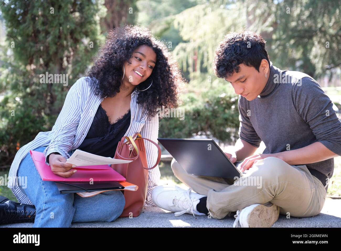 Two latin students smiling studying together sitting on a bench outdoors. University life at campus. Stock Photo