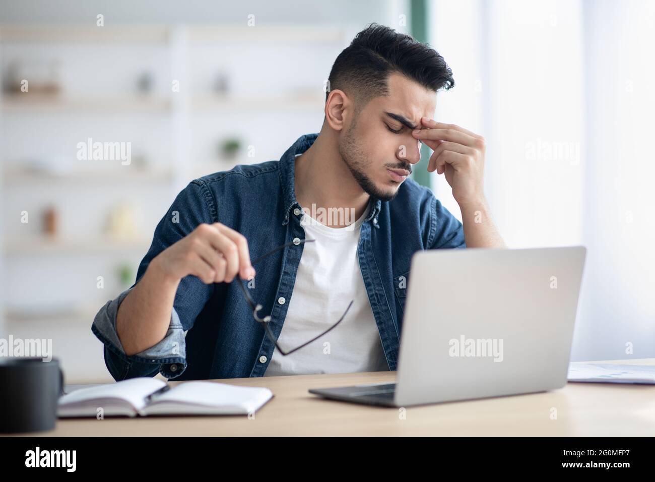 Exhausted arab guy sitting at workdesk, removing glasses Stock Photo