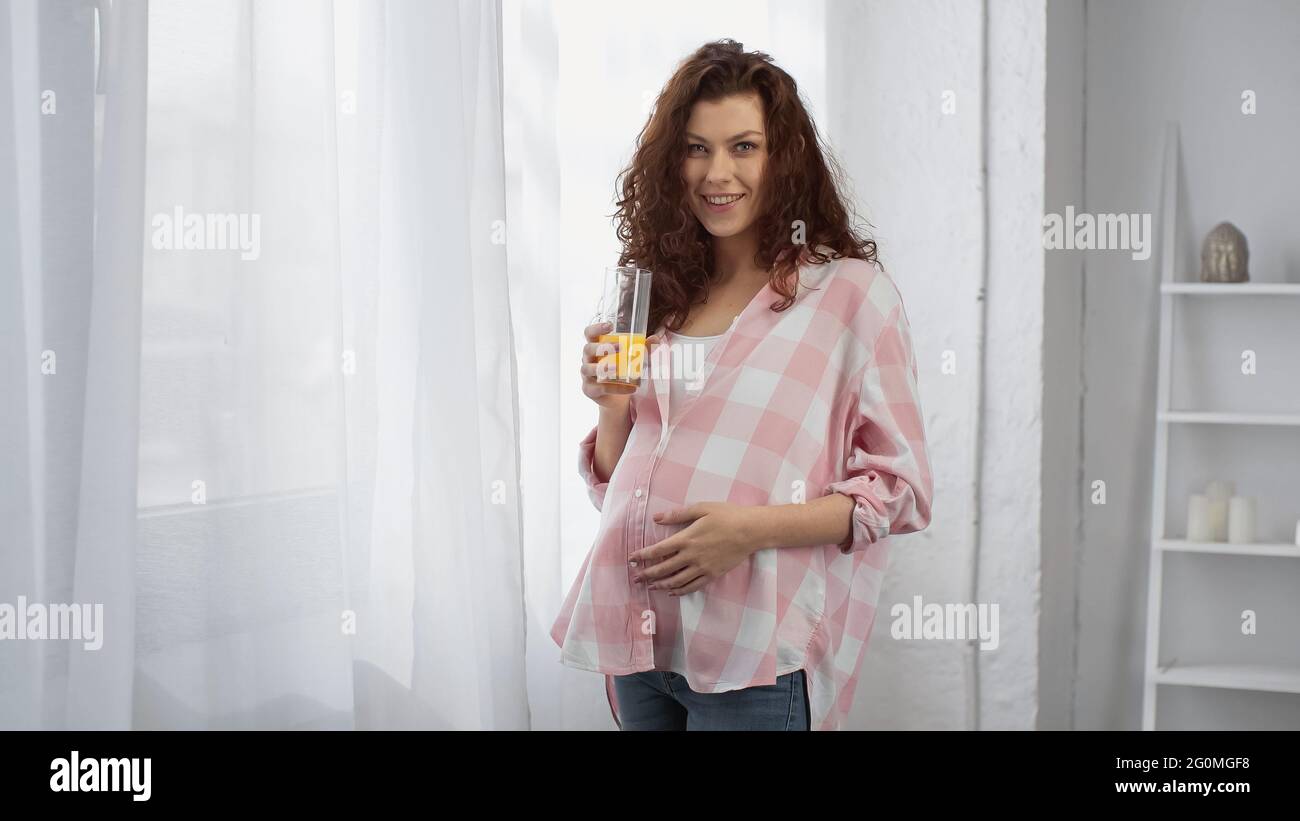 pregnant and curly woman smiling while holding glass of orange juice ...