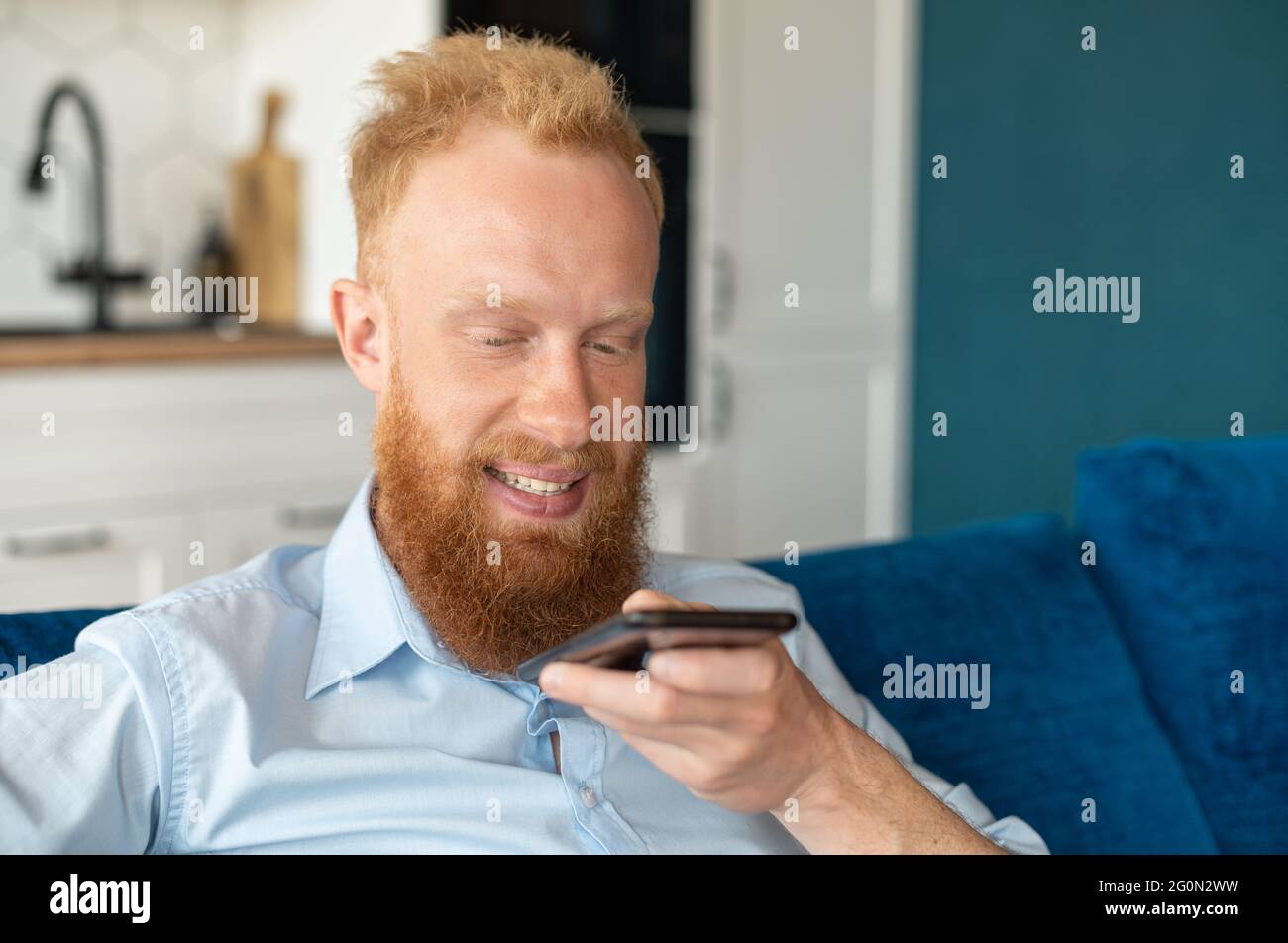 Charismatic redhead guy record voice message and smiling, sits on the sofa at home and holding smartphone near mouth and talking in the mic, chatting, using voice recognizable mobile application Stock Photo