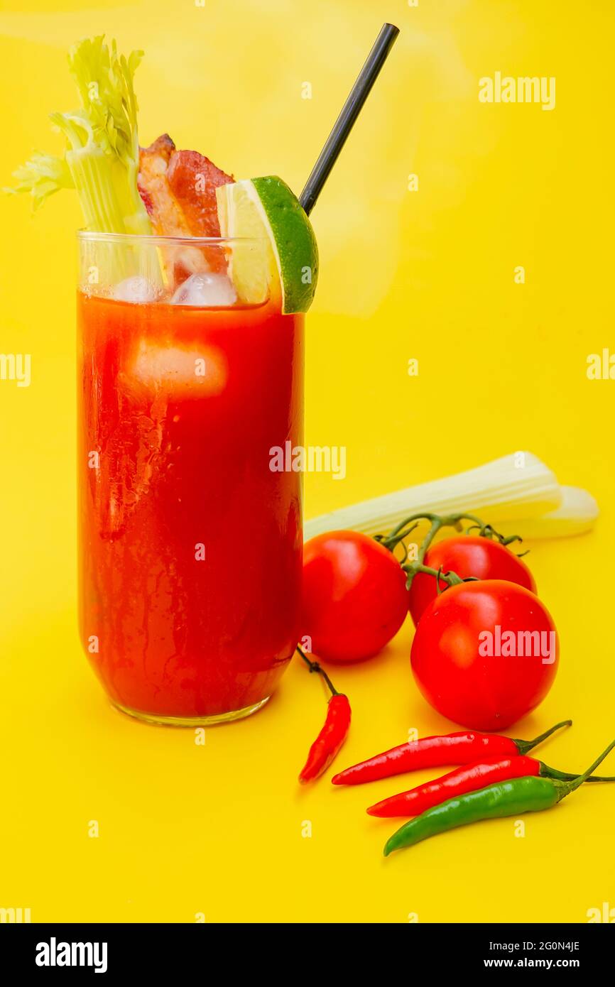 Bloody Mary in a glass with a straw, celery , lime ,bacon and ice. On a yellow background with fresh vine tomatoes, celery and fresh cayenne peppers . Stock Photo