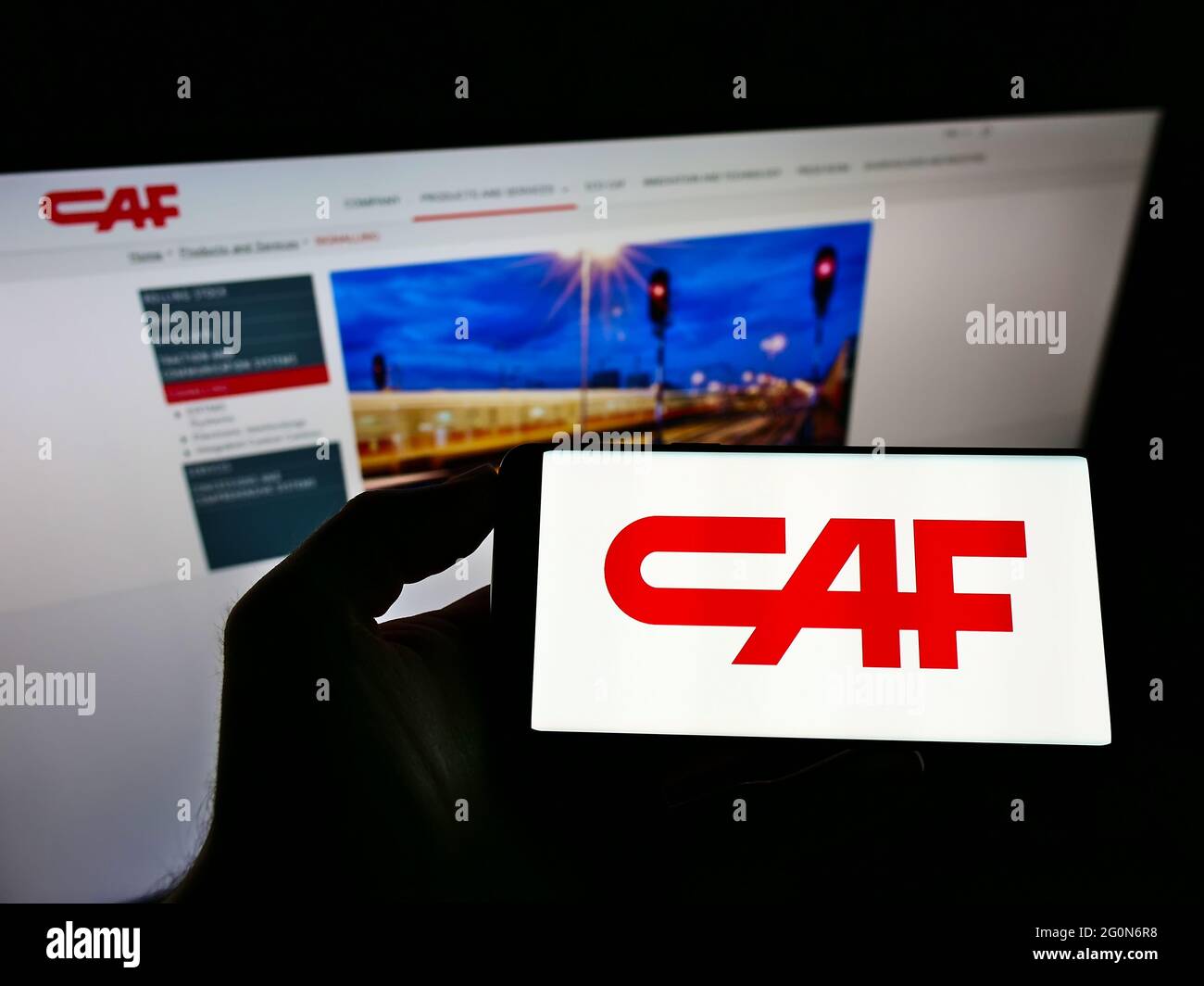 Person holding cellphone with logo of Construcciones y Auxiliar de Ferrocarriles S.A. (CAF) on screen in front of web page. Focus on phone display. Stock Photo