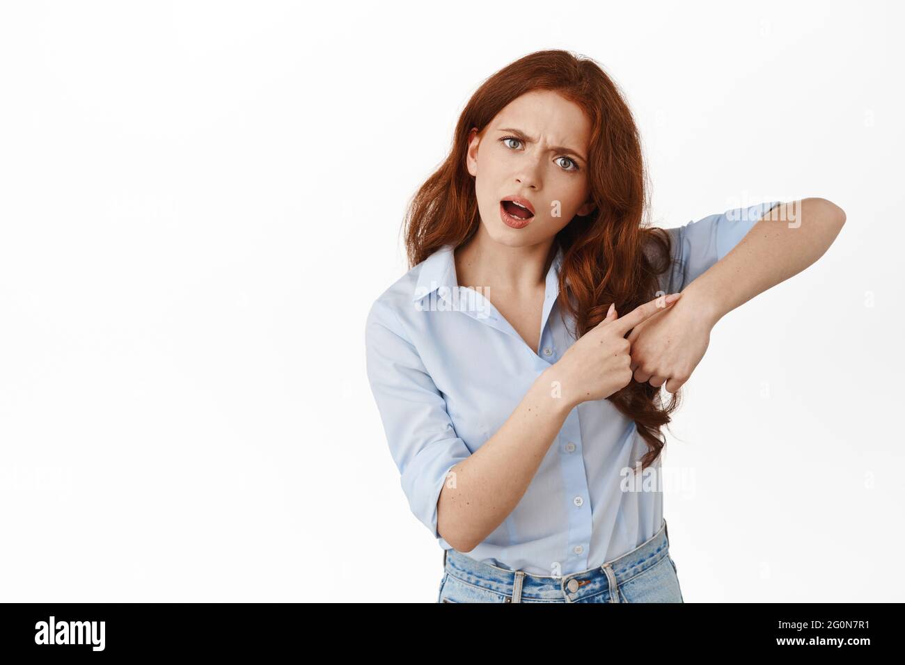 Hurry up. Annoyed redhead girl pointing at watch wrist gesture, telling to move faster, we are late, telling the time, standing in blouse against Stock Photo