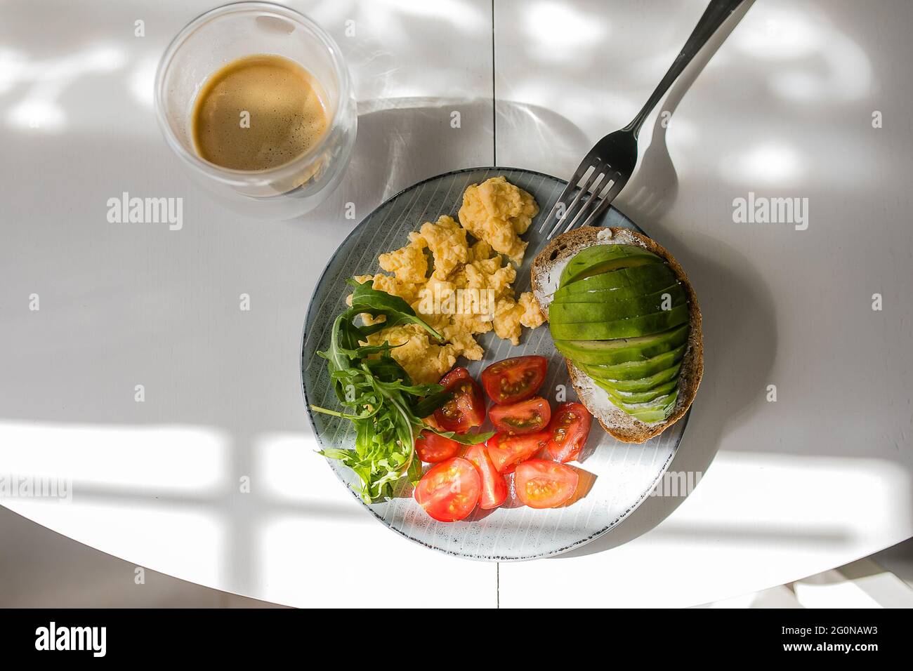 Plate with scrambled eggs, arugula, tomato, whole wheat toast with avocado, cup of coffee on table. Stock Photo