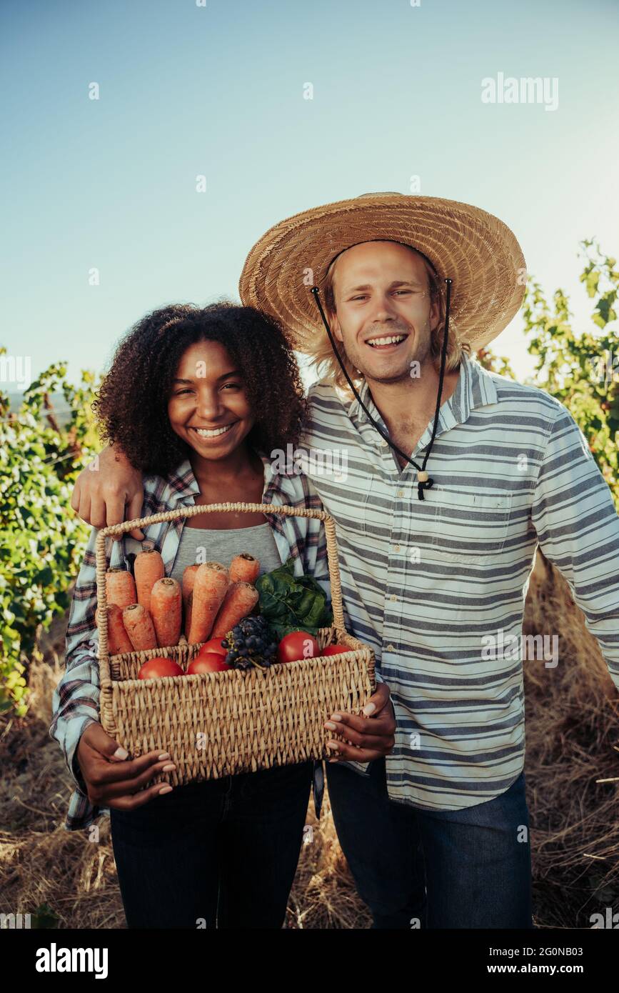 Mixed race farmers laughing and smiling after long day of harvest holding full basket of fresh vegetables  Stock Photo