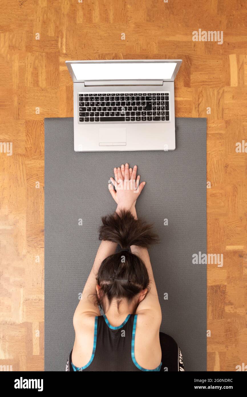 Unrecognizable woman with tied hair doing yoga on the floor while using a computer to teach online classes. Stock Photo