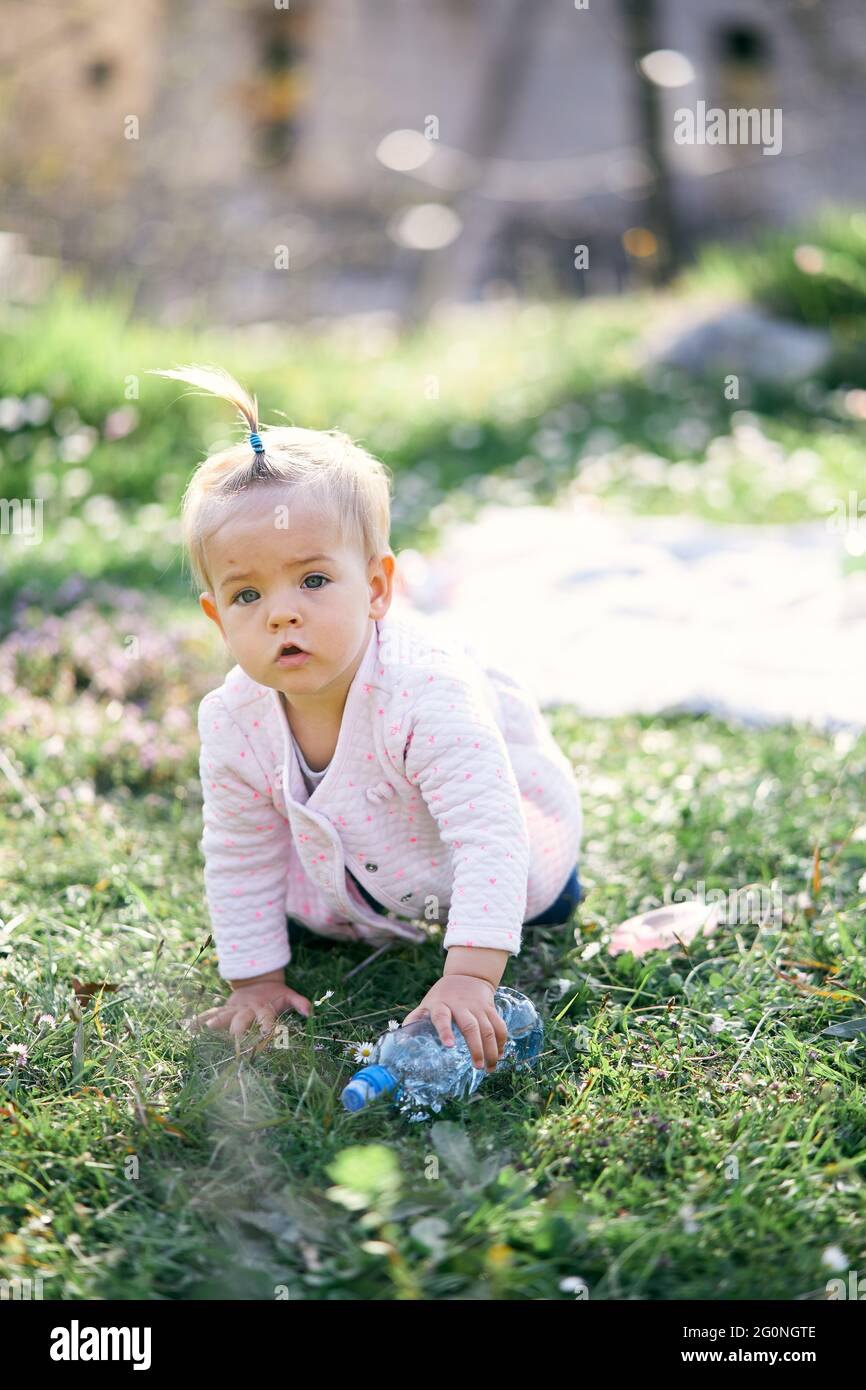 Pensive little girl with a ponytail on her head crawls along a green lawn among flowers and holds a plastic bottle in her hand Stock Photo