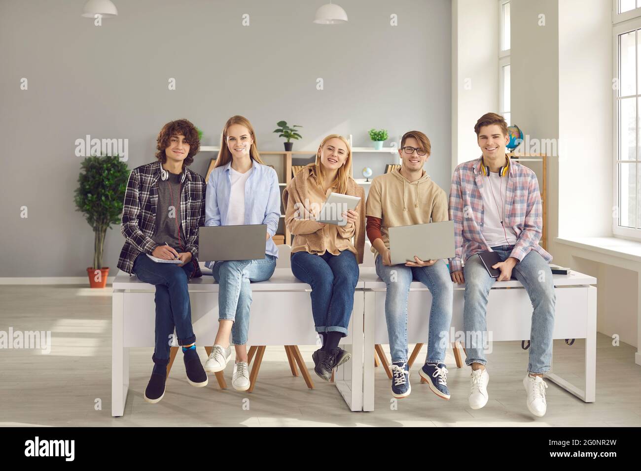 Group of happy school, college or university students sitting on desk with digital devices Stock Photo