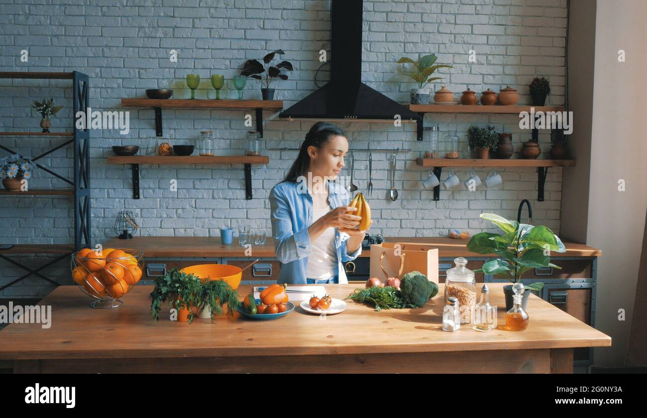 A beautiful young woman in a good mood is sorting vegetables from a bag and putting them on the kitchen table.Happy young housewife lays out purchases Stock Photo