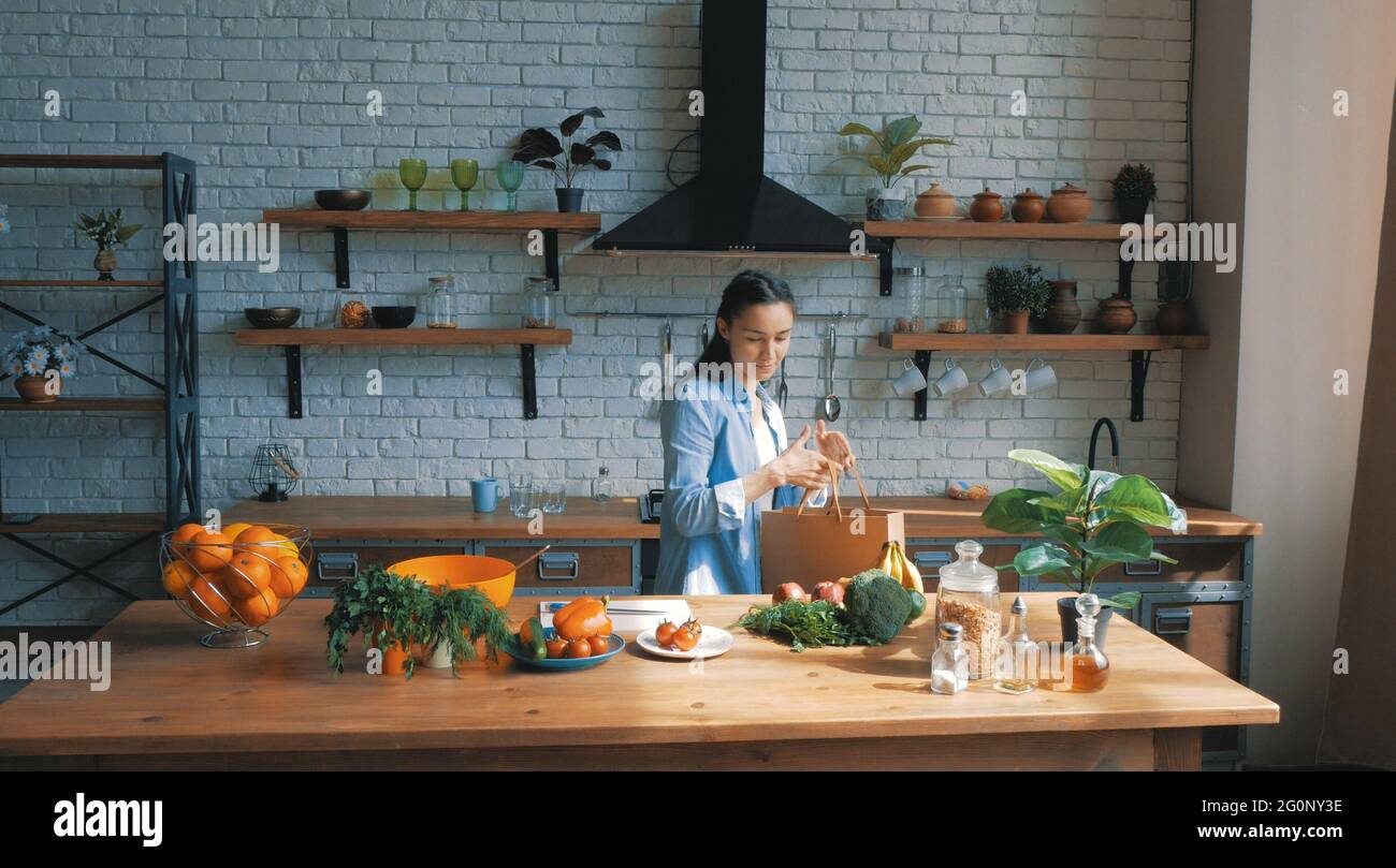 A beautiful young woman in a good mood is sorting vegetables from a bag and putting them on the kitchen table.Happy young housewife lays out purchases Stock Photo