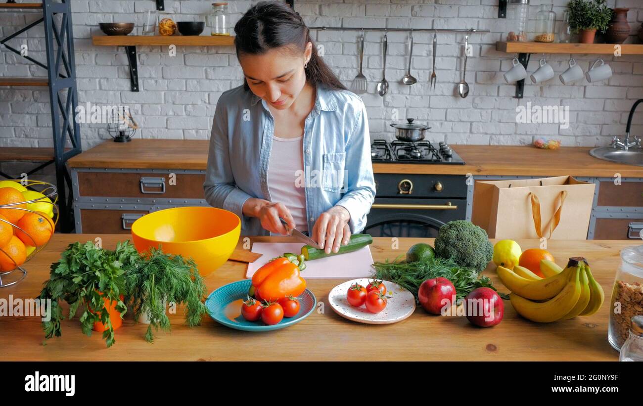 A beautiful young woman in a good mood cuts a cucumber for a salad. Happy young housewife prepares vegetable salad in her kitchen. Young beautiful Stock Photo