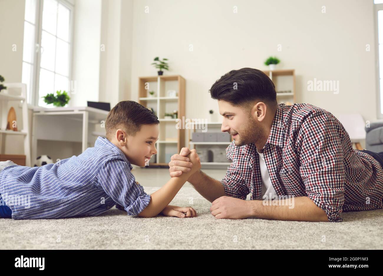 Side view of happy father and son competing in arm wrestling lying on the floor at home. Stock Photo