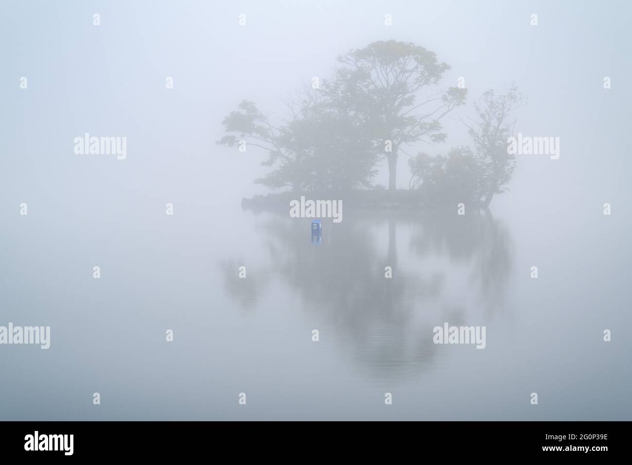 The small island on Yeadon Tarn (dam) is isolated by thick fog on a late spring morning, creating a minimalist scene reflected in the calm water. Stock Photo