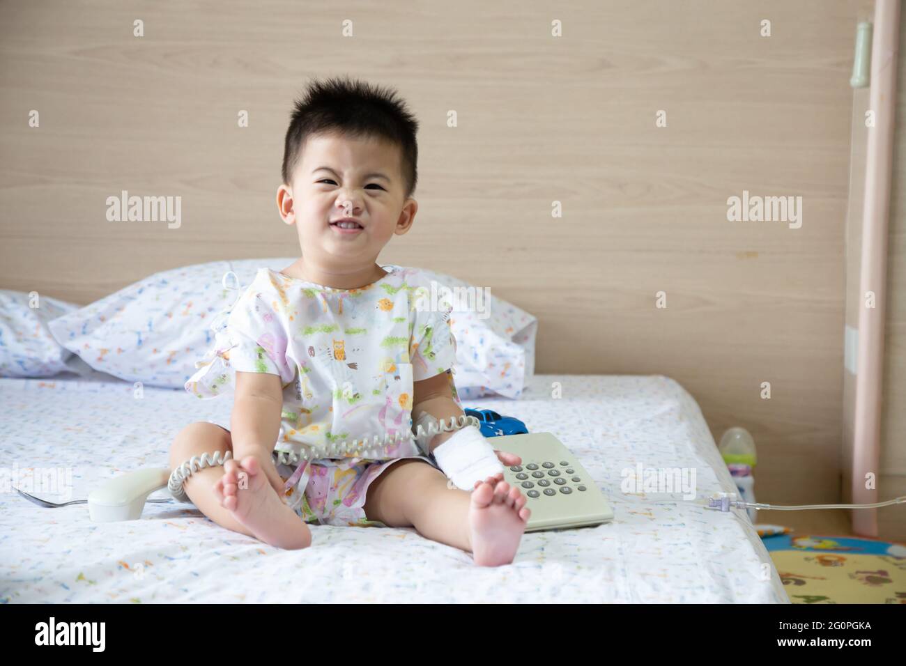 Asian baby boy smiling and wear patients suits sitting on bed in the room at child department in the hospital. Children with infectious diseases IPD, Stock Photo