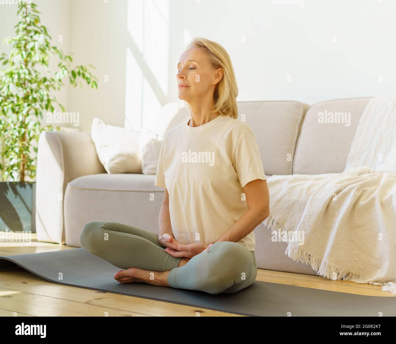 Peaceful senior woman in lotus position meditation with closed eyes at home while sitting on yoga mat on floor, full length. Calm elderly lady practicing meditation techniques and yoga indoors Stock Photo