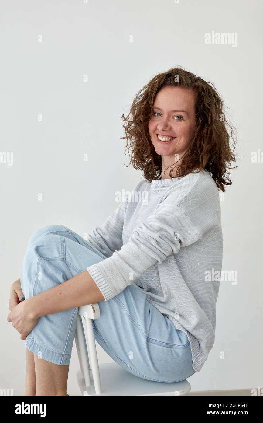 Side view of young female in trendy outfit sitting on wooden chair on white background and looking at camera Stock Photo