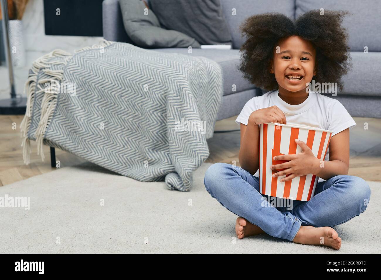 Positive child holds large popcorn bucket while watching a cartoon movie at home cinema. African American little girl is sitting on home floor, eating Stock Photo